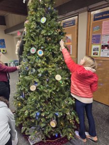 A blonde girl in a red jacket reaches to place an ornament on the artificial Christmas tree in Sandburg Hall