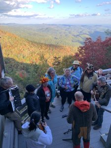 A group of 11 people gathered on a deck overlooking an expansive mountain range. Some people are holding musical instruments, some are wearing costumes. 